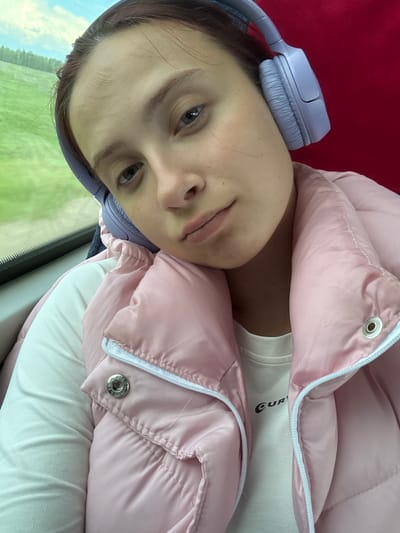 A young woman is pictured wearing lilac headphones and a pink puffer jacket, with a white t-shirt visible beneath the jacket. She appears to be inside a vehicle, possibly a train, as suggested by the blurred green landscape visible through a window in the background. There are no explicit cues in the image to confirm the location as Kukushtan, Russia.