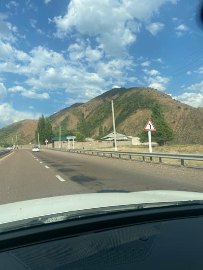 The image captures a view from inside a vehicle, looking out at a road flanked by a metal guardrail. A white car is visible ahead on the right lane of the road, which appears to be a multi-lane highway. The road curves to the left as it approaches a settlement. On the right side of the road, a signpost with a triangular warning symbol indicates a sharp left turn. Further ahead, a sign reads "KOKSAROY," suggesting this is the location. Several trees, including tall, slender ones and some evergreen trees, dot the landscape. Behind the settlement, a brown, rugged mountain range rises, partially covered with vegetation. The sky above is blue with scattered white clouds. The overall scene suggests a scenic drive through a mountainous region in or near Beshkul, Uzbekistan.