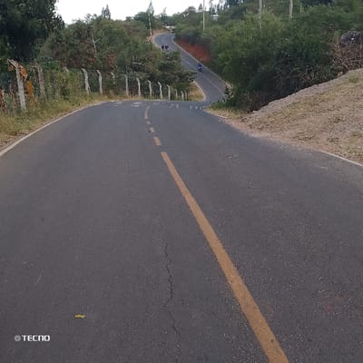 A winding paved road slopes downward with a solid yellow line painted near the center. Green vegetation lines both sides, punctuated by a fence on the left. Further up the road, people walk along the roadside, and a person rides a motorcycle. The sky is overcast. A crack runs through the road surface in the foreground. A "TECNO" watermark is present in the lower left corner.