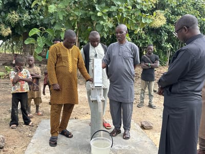 In Afogo, Nigeria, a group of seven individuals, including four men and three children, are gathered around a water pump. The men, dressed in traditional African attire and casual clothing, appear to be observing or operating the pump, which is situated on a concrete base. A white bucket filled with water sits at the base of the pump. The background consists of lush green foliage and trees, suggesting an outdoor, rural setting. The ground is covered in soil and scattered rocks.
