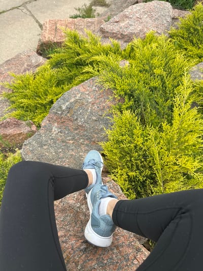 A close-up, eye-level shot depicts a person standing on a rock formation adjacent to a patch of vibrant green shrubs. The individual is wearing black leggings, white socks, and light blue and white athletic shoes. The foreground includes the person's legs and shoes, which rest on a reddish-brown granite rock. To the left, a dense cluster of yellowish-green juniper or cypress shrubs spills over another rock. A paved walkway can be seen in the distance, to the bottom left of the image. The image shows a corner with some concrete and the edge of green grass on the left, the center with a rock formation that is covered in juniper or cypress shrubs and the feet of a person in leggings and sneakers standing on the rock formation. Zakharivka, Ukraine is not discernible from the image.