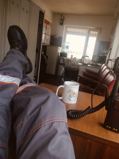 The image depicts an indoor scene, possibly an office or break room, with elements suggesting a location in Salsk, Russia, though no direct visual cues confirm this. In the foreground, a person's legs are prominently displayed, clad in dark grey work trousers with reflective orange stripes near the ankles. They are wearing black work boots. The legs are propped up on what appears to be a wooden desk.
On the desk, a white ceramic mug with a design of a horse is visible. Next to it, a black coiled cord leads to a black electronic device, likely a radio or walkie-talkie, which is also on the desk.
The background reveals a room with lockers on the left, marked with lines. Further into the room, a window with natural light streaming in is a focal point, showing a glimpse of the outdoors. Adjacent to the window are various appliances and furniture, including what looks like a water cooler, a kettle, and chairs stacked on top of each other. The overall impression is of a functional, utilitarian space.