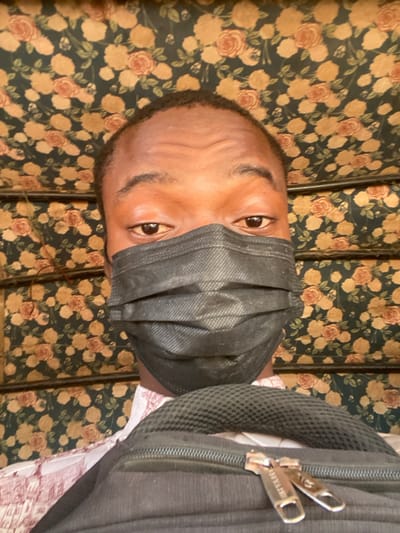A person wearing a black face mask is looking at the camera with a backpack strapped on their right shoulder. The background is a busy floral pattern in shades of brown, tan, and green. The person's skin tone is brown, and their eyes are visible above the mask. The photo seems to be taken indoors. The backpack is black and partially obscures the person's body.