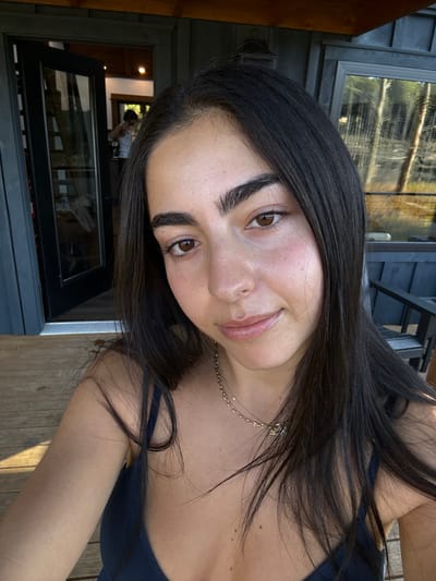 A young adult female is centered, positioned outdoors on a light brown wooden deck, likely in Otego, United States. She has long, dark hair, brown eyes, full eyebrows, and wears a dark-colored, thin-strapped top and a delicate gold chain necklace. She looks directly at the camera with a slight smile. To her left, a dark-colored door with multiple glass panes is ajar, revealing an out-of-focus individual, appearing male and wearing a light tank top, standing inside a room with wooden walls. To her right, a dark exterior wall with horizontal siding contains a window reflecting tall trees. A dark, slatted outdoor chair is partially visible on the right. The scene is illuminated by natural daylight.