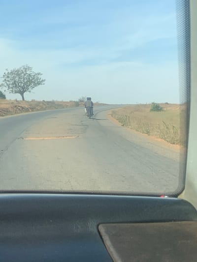 A motorcyclist is seen in the distance on a paved road, likely viewed from inside a vehicle. The road curves slightly and is bordered by dry vegetation. A lone tree stands to the left of the road. The sky is pale blue with a hint of cloud cover. The interior of the car is visible on the right side of the frame.