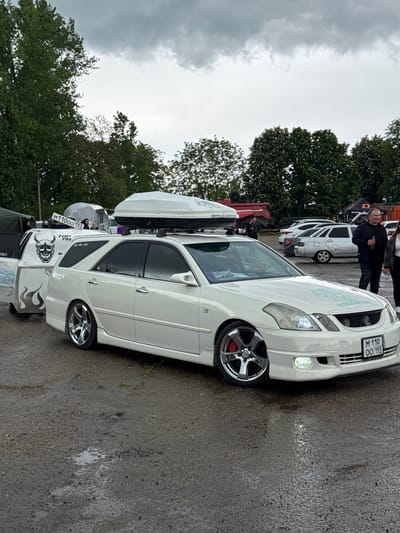 A white station wagon with custom wheels and a roof-mounted cargo carrier is parked on a wet asphalt surface. The vehicle features decorative graphics on the rear quarter panel and custom grill work. The license plate reads "M 110 OO 193". Several other vehicles are visible in the background, along with people. The location is Pavlovskaya, Russia, as indicated by the license plate code. Trees are in the background, and the sky is overcast. A man in dark clothing is standing behind the car.