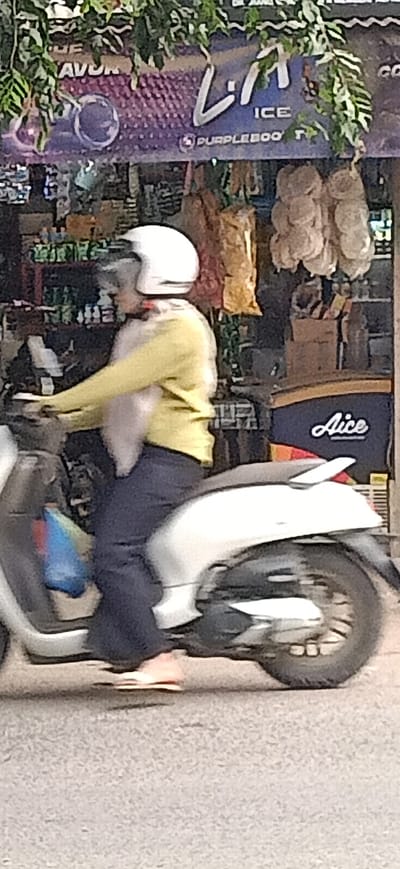 A person wearing a white helmet and a yellow long-sleeved shirt is riding a white scooter on a street in Gampongteungoh, Indonesia. The person is wearing dark blue pants and pink sandals. A blue and red bag is visible on the left side of the scooter. In the background, there is a shop with a sign that reads "LI ICE" and "PURPLEBOO T". There are also hanging items that look like dried goods and some bottles on shelves. The scene appears to be outdoors during the day. The lighting suggests it might be sunny. The overall impression is a casual street scene in a town.