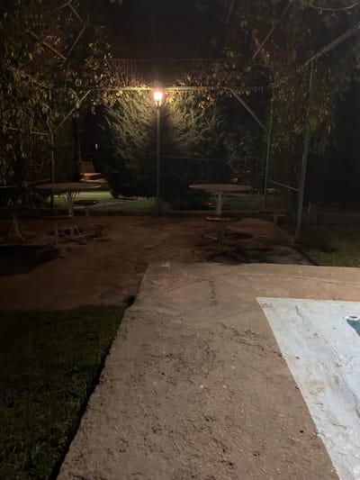 This image depicts an outdoor seating area at night, likely in the city of Mashat, Kazakhstan. The scene is illuminated by a single lamp post, casting a warm glow on the surrounding greenery and paved surfaces. Several round tables with accompanying stools are arranged under a covered structure, suggesting a place for dining or relaxation. The ground is a mix of concrete and grass, with a pool edge visible in the lower right corner, hinting at a recreational facility. The overall atmosphere is tranquil and slightly mysterious, with the darkness of night enveloping the scene.