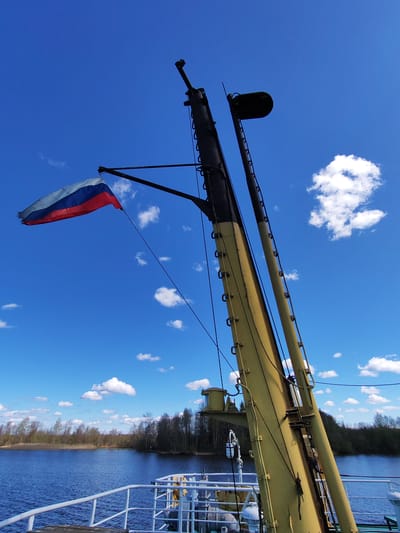 A low-angle, outdoor shot shows the upper portion of a boat's mast against a blue sky dotted with white clouds. The mast is painted yellow and black, and features a Russian flag fluttering in the breeze. In the background is a body of water with wooded shores. The location is likely in Chaika, Russia, based on the presence of the Russian flag and general landscape. There are no people visible in the image.