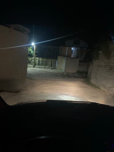 An interior shot from the driver's perspective shows a dark road at night, illuminated by headlights. The road appears to be paved with dirt or gravel and is somewhat uneven. On the left side of the road is a light-colored building with a textured wall. Further down the road, on the right, is a rough, stone wall. In the background, there are structures that appear to be houses or outbuildings, some with visible windows. A street light is visible on the left, casting a bright glow. Trees and shrubbery are present in the background, especially to the left of the road, near the street light. The overall scene suggests a rural or semi-rural setting within the city of Gergebil, Russia, at night.