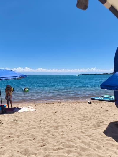 A sunny day at the beach in Bayet, Kyrgyzstan, is captured in this image. The foreground is dominated by sand, with a few flip-flops and a towel suggesting recent activity. A blue umbrella provides shade to a person wearing a straw hat and a colorful patterned dress, standing near the water's edge. To their left, a blue bag and orange flotation device are visible. In the water, a person is visible in a green flotation ring. Further out, the clear blue water meets a distant horizon line where scattered white clouds hover above a landmass. Several paddleboards are arranged on the sand to the right of the frame, with two blue umbrellas also present. The overall scene depicts a leisurely day at the lake.