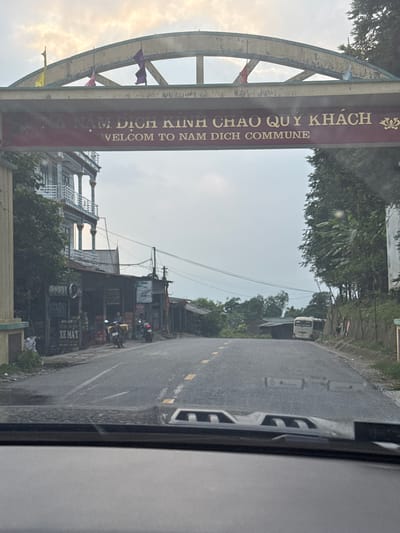 The image captures a roadside scene, appearing to be taken from inside a moving vehicle, looking forward. The camera is held vertically, presenting the scene rotated 90 degrees clockwise.

In the foreground and central to the image is a large, ornate archway spanning over a paved road. The archway has a maroon horizontal banner with golden Vietnamese text that reads "NAM DỊCH KINH CHÀO QUÝ KHÁCH" and below it in English, "WELCOME TO NAM DICH COMMUNE". Various small, colorful flags (purple, pink/red, yellow) are strung across the top of the archway's metallic framework.

The road ahead is asphalt, with white and yellow dashed lane markings. To the right, the side of a vehicle's dashboard or door frame is visible. Further down the road on the right, there are several buildings, likely shops or homes, with various signs. One sign clearly displays "XE MÁY" (Vietnamese for "motorbike") and what appears to be a phone number. Several motorbikes are parked or moving near these buildings. Power lines crisscross the sky above the road.

On the left side of the road, more buildings and dense green foliage from trees are visible, extending into the background. A light-colored bus or truck can be seen further down the road on the left. The sky is bright but appears overcast or hazy, suggesting it's daytime, possibly late afternoon or early morning. The overall setting depicts an entrance to a rural or semi-urban commune in Vietnam.