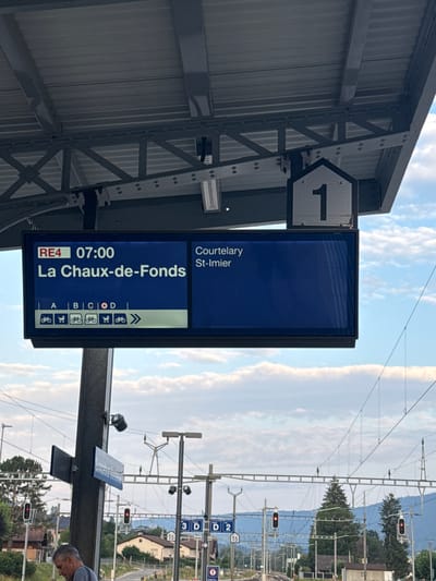 The image shows a train station platform in Sonceboz-Sombeval, Switzerland. A digital display board indicates that train RE4, departing at 07:00, is bound for La Chaux-de-Fonds and will stop at Courtelary and St-Imier. Below the destination information, symbols indicate available spaces for bicycles and wheelchairs on different train sections labeled A, B, C, and D. The platform roof is supported by a metal structure. In the background, railway tracks, overhead electrical lines, traffic lights, and buildings can be seen. A man is partially visible on the left side of the image. The sky is cloudy.