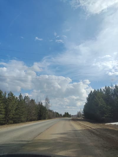 The image presents a daytime view from the perspective of a vehicle traveling or stopped on a rural road in a forested area, consistent with the landscape found in Krasnoyar, Russia. The road, appearing to be paved with asphalt that transitions into a wider, unpaved dirt or gravel shoulder, extends straight into the distance before gently curving.
Dense coniferous forests, primarily composed of pine trees, line both sides of the road. On the left side and further into the distance on the right, some bare deciduous trees, likely birches, are interspersed among the evergreens, suggesting the season is late winter or early spring. A small patch of snow is visible on the dirt shoulder on the far right, reinforcing the seasonal inference.
The sky is a prominent feature, a clear and bright blue, dotted with fluffy white cumulus clouds that stretch across the expanse. Several thin, dark power lines run horizontally across the upper half of the image, cutting across the sky above the tree line. No people or other vehicles are visible, contributing to a sense of solitude and vast natural landscape. The weather appears sunny and pleasant. There is no visible text in the image.