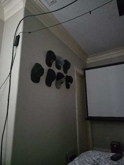 The image shows a living room in a house located in Happy Valley, Oregon, at the coordinates 45.447 latitude and -122.544 longitude. The room is sparsely furnished, with only a bed and a projector screen visible. There is a projector on the floor next to the bed, and a remote control is on the bed. The wall is painted gray, and there are eight black baseball caps hanging on the wall. Black electrical cords are attached to the ceiling. The room is dimly lit, and it appears that no one is present.