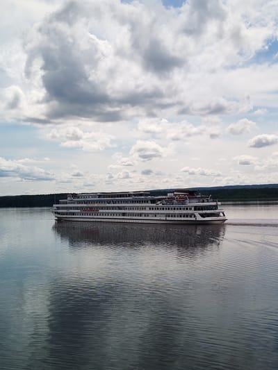 A large white multi-deck riverboat cruises on a calm river under a cloudy sky. The riverboat, with multiple windows and decks, is seen from its port side, moving from left to right across the frame. A red banner with Russian text is visible on the side of the boat. The river reflects the boat and the sky, with gentle ripples on the water's surface. In the background, a dense green forest lines the opposite bank of the river. The sky is filled with dramatic, voluminous white and grey clouds, with patches of blue visible. The overall scene is one of serene travel on a river, likely in a natural setting. The location of Shchetinino, Russia, is mentioned, suggesting this is a scene from that area.