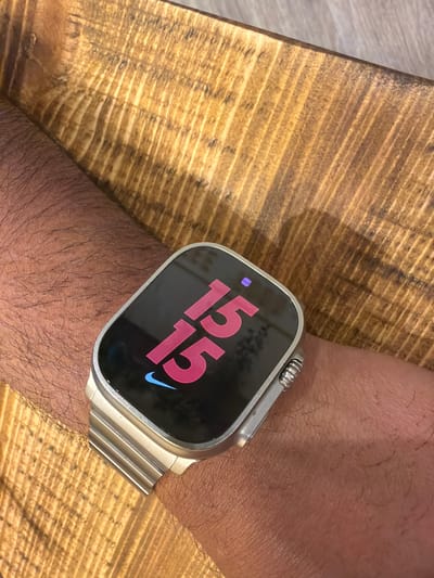 A close-up shot shows a wrist wearing a smartwatch, possibly an Apple Watch Ultra with a silver metal link bracelet. The watch face displays "15 15" in a pinkish-red hue, accompanied by a blue Nike logo. The arm is hairy and appears to be resting on a wooden surface. No specific location in Mole Valley, United Kingdom is discernible from the provided image, as the scene is too close-up and lacks contextual clues.
