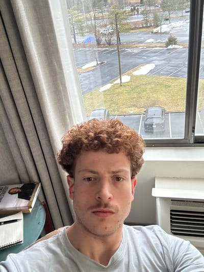 An indoor scene captured from a high vantage point looks out onto an outdoor environment.
**Indoor Environment:**
A fair-skinned male with curly reddish-brown hair, a short mustache, and light stubble is centered in the foreground, wearing a light gray t-shirt and looking directly at the camera. To the left, light beige textured curtains are partially drawn. Below the left side of the window, a light-colored table is visible, holding a spiral-bound notebook with a black pen and a stack of books, one displaying a person's image and text, possibly "TRADING STRATEGY." To the right, below the window, a white air conditioning unit is mounted.
**Outdoor Environment (View through window, located in Fairfield, United States of America):**
The window overlooks a large asphalt parking lot with numerous marked spaces, many of which are empty. Patches of melting snow are scattered across the parking lot and adjacent grassy areas, indicating a cold season. Green grass is visible where snow has melted. Two dark-colored vehicles are parked below the window frame in the immediate foreground of the lot. Several bare deciduous trees and some evergreen shrubs are dispersed throughout the scene. A tall, dark utility pole stands prominently in the middle of the parking lot view. In the distance to the left, a bright blue tarp is visible among trees. Further background elements include additional buildings and distant trees, suggesting a suburban or commercial area.