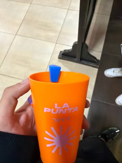A hand holds an orange cup with the words "La Punta Wet" and a pink sunburst design. A blue ice pop protrudes from the cup. The person wearing black pants and black and green shoes is standing on gray and beige tiled flooring. A black table leg is visible in the left of the frame. The image suggests a location where La Punta Wet products might be available, perhaps Punta Umbría, Spain.