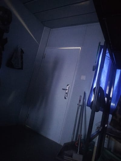 This media file depicts an interior scene, likely a room or hallway, with a door and various objects. The lighting is dim, with a bright light source emanating from the top of the door, casting a strong glare. On the left side of the image, a shadowy figure or object is visible against the wall. A white bag hangs on the wall.
In the foreground and to the right of the door, there are cleaning tools, including what appear to be mops or brooms. Behind these, blue curtains are visible, partially obscuring shelves or a rack. A metal structure, possibly part of a bunk bed or shelving unit, is also present.
The overall impression is one of a dimly lit, somewhat cluttered space. The time of day is not explicitly indicated, but the low light suggests it could be evening or night, or a space with limited natural light. There are no clear weather cues. Location cues are minimal, with the provided context being "Калыс-Ордо жаңы курулушу, Kyrgyzstan". There is no visible text in the image. The scene appears to be static, with no active event taking place. The emotion conveyed is ambiguous due to the low light and lack of clear subjects, but the stark lighting might suggest a sense of mystery or unease.