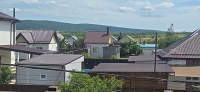 This media file depicts a rural residential scene in Gostagayevskaya, Russia, on a cloudy day. The foreground is dominated by the roofs of several houses, constructed with brown and grey corrugated metal. One house on the far left has a white exterior with dark trim and a flat, extended brown roof. Adjacent to it is a house with a pitched brown roof and white siding with dark trim. Further into the scene, other houses are visible, some with white exteriors and pitched roofs of varying colors, including brown and grey. There's also a smaller, wooden shed-like structure with a simple roof.
The middle ground shows a fence, possibly a garden or yard, and more greenery from trees and bushes. Beyond the houses and fences, a gently rolling landscape stretches out, covered in green fields and scattered trees. In the distance, a large, tree-covered hill or mountain range forms the backdrop under a pale blue sky with scattered white clouds. The overall impression is one of a quiet, suburban or rural settlement. There are no people visible in the image, and the scene appears to be static, with no discernible activity. The time of day is likely daytime, given the brightness of the scene, and the weather is overcast. No visible text is present in the image.