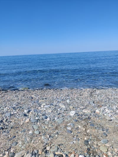 The image depicts a pebbled beach meeting the tranquil, clear blue sea under a bright, cloudless sky. The foreground is dominated by a dense scattering of smooth, varied-colored pebbles and sand, with some larger stones visible near the water's edge. The water itself is calm, with gentle ripples and a translucent quality that allows glimpses of the submerged pebbles. In the distance, a faint, hazy landmass is visible on the horizon, a subtle line separating the sea and sky. The scene is devoid of any people, structures, or human activity, presenting a natural coastal environment. The location is identified as a beach within the municipal unit of Evrymenon, Greece.