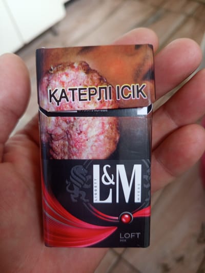 A close-up, eye-level shot shows a hand holding a pack of L&M Loft Mix cigarettes. The pack is black with red and white accents. A graphic image of a cancerous lung is visible on the front of the pack, with the words "ҚАТЕРЛІ ІСІК" (meaning "Malignant Tumor" in Kazakh) printed in white over the image. The brand name "L&M" is displayed prominently in large white letters, with smaller text that reads "LIGGETT MYERS". Below this, a red circular graphic element is present, followed by "LOFT MIX" in white text. The background is blurred, suggesting an indoor environment. No specific details about the location in Кенжекөл, Kazakhstan are discernible from the image.