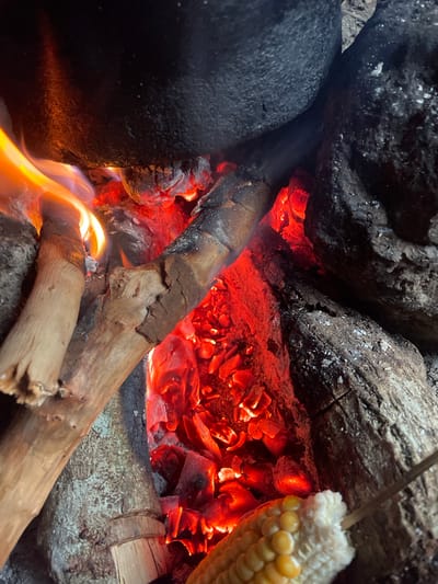 This image captures a close-up view of a campfire with glowing embers and flames. The scene is set outdoors, likely in Aiyegunle, Nigeria, given the location context. The primary subjects are the burning wood, coals, and a stick with a cob of corn skewered on it, positioned to be roasted over the fire.

The fire is the central focus, with vibrant red and orange coals radiating heat and light. Pieces of burnt wood are scattered around, some still smoldering. The flames are visible licking at the lower part of the frame, adding to the dynamic and warm atmosphere. The corn on the cob appears to be nearing completion, with some kernels already showing signs of charring.

The background consists of large, dark rocks and more burnt wood, suggesting a rustic or natural setting. The textures of the rocks and wood are rough and weathered. There is no visible text in the image. The time of day is not explicitly indicated, but the intensity of the fire suggests it might be evening or nighttime, or a shaded area where the fire's glow is prominent. The overall mood is one of cooking and perhaps relaxation around a campfire.