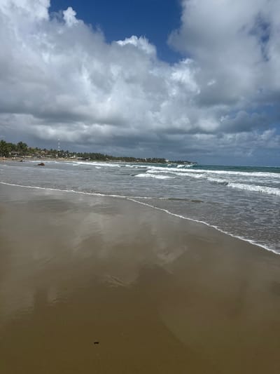 The image shows a beach scene in Cabarete, Dominican Republic. The sky is partly cloudy with a mix of blue sky and white and dark gray clouds. The beach is wide with tan sand. Waves are visible as they roll onto the shore. Along the shoreline, there are palm trees and buildings, including what appears to be a tall white tower. The overall impression is a beach on a cloudy day.