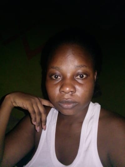 The image displays a close-up, head-and-shoulders portrait of an adult female, positioned centrally and facing directly towards the camera. The subject exhibits dark skin and dark hair. She is wearing a plain white sleeveless tank top. Her right hand is raised, with fingers gently curled and resting against her lower jawline on the viewer's left side. Her eyes are open and directed straight ahead, maintaining a neutral facial expression. A small, dark, raised mark is visible on her upper lip, situated slightly to the viewer's right of the philtrum. The background consists of a dark olive-green wall, dimly lit, suggesting an indoor environment. A faint, angular, dark red line, possibly a decorative element or structural feature, is visible in the upper left background. No specific visual identifiers within the image confirm its location in Sabongidda Ora, Nigeria.