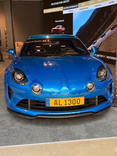 The image shows a bright blue Alpine sports car parked indoors on a grey carpet. The car is positioned centrally and faces the viewer. On the car's windshield, text reads "Drive from 99 €". The license plate is yellow with black lettering, reading "AL 1300", and features the Luxembourg flag emblem on the left. In the background, a large screen displays driving experiences, showing text like "PILOTAGE SUR CIRCUIT", "DRIVING EXPERIENCES", "FERRARI", "LAMBORGHINI", "DRIFT", "MOTO", "KIDS", "PORSCHE", and "ALPINE". The screen also shows images of cars on a race track and text that mentions "Ferrari 488 GTB" and "HOCKENHEIM". The environment appears to be a showroom or exhibition space in Reckange-sur-Mess, Luxembourg. There are no people visible in the frame.