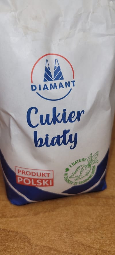 This is a close-up, rotated image of a white paper bag, likely containing sugar. The bag is on a textured, brown surface, possibly a table. The dominant text on the bag is in Polish. Large, cursive blue lettering spells out "Cukier biały," which translates to "white sugar."
To the left of this text is a circular logo with a red border. Inside, in blue lettering, is the word "DIAMANT," the brand name. A stylized blue and white logo, resembling a mountain or abstract sail, is positioned above "DIAMANT."
To the right of "Cukier biały" is a green graphic of a carrot with leaves, enclosed in a circle. Below this graphic, also in a circular arrangement, is text that reads "W ZGODZIE ZE ŚRODOWISKIEM," meaning "In harmony with the environment."
Towards the bottom right of the bag, there is a red and white rectangular label with blue text. The top part reads "PRODUKT" and the bottom part reads "POLSKI," indicating "Polish product." The bag's design features blue accents along its edges. The lighting suggests an indoor setting, and the overall impression is that of a product package.
