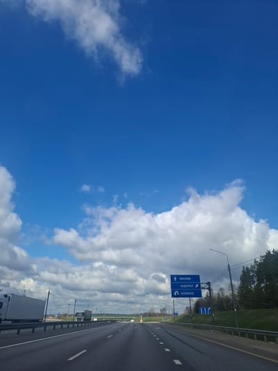 This image captures a highway scene under a bright, partly cloudy sky. The perspective is from a vehicle traveling on the multi-lane road, which stretches into the distance. White dashed and solid lines mark the lanes, and a metal guardrail runs along the right side. Beyond the guardrail, there's a grassy shoulder followed by a line of trees or dense foliage. The background reveals a flat, open landscape beneath the horizon.
A prominent blue road sign is visible on the right side of the highway, supported by a metal gantry structure. Streetlights with extended arms are spaced along the roadside. Parts of large vehicles, including the front of a white truck in the foreground and another truck further down the road, indicate ongoing traffic.
The weather is clear daytime, with vivid blue sky dominating the left half of the image and an expansive bank of fluffy white clouds occupying the right.
The most notable detail is the text on the blue road sign, which is in Cyrillic:
- An upward arrow points towards "МОСКВА" (MOSKVA / MOSCOW).
- Another upward arrow points towards "ЗАДОНСК" (ZADONSK).
- A U-turn arrow points towards "ХЛЕВНОЕ" (KHLEVNOYE).
A smaller blue sign further down the road displays multiple upward arrows, likely indicating straight-ahead lanes. The overall scene suggests travel on a highway in Russia, consistent with the location context of Staraya Voskresenovka.