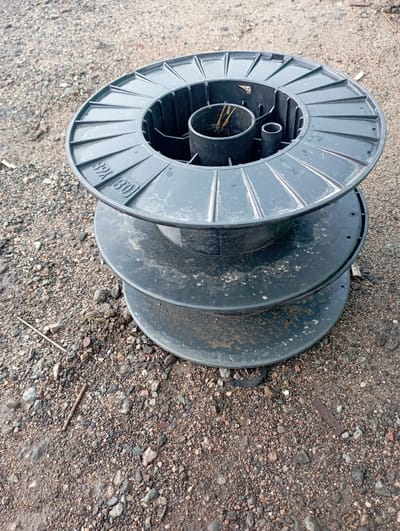 The image shows a stacked set of three black plastic spools, possibly wire or cable spools, resting on a surface of gravel and dirt. The spools are empty and appear weathered, with some dirt and debris on their surfaces. The top spool is the most detailed, with visible ribbing on its outer edge and markings that include "32k" and "(80)". The background consists of a rough, granular ground surface. The location is identified as Бәйтерек, Kazakhstan. There are no people or animals visible in the image.
