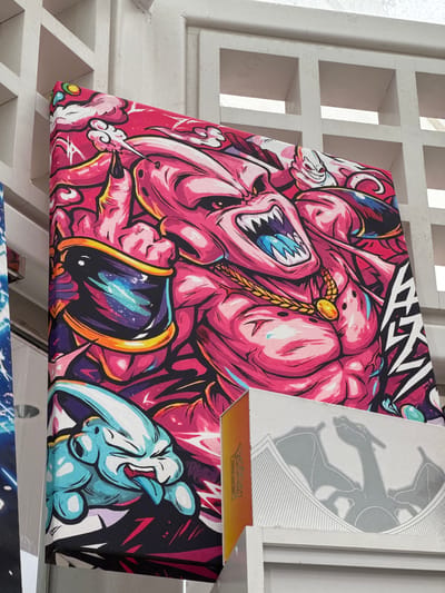 The image shows a close-up of a vibrant, stylized artwork featuring characters from the Dragon Ball Z anime. The main subject is a large, pink, muscular character with an aggressive expression, sharp teeth, and yellow jewelry. To the bottom left, a smaller, light blue character with a similar aggressive expression is shown sticking out its tongue. The artwork is printed on a canvas that is propped up against a white, lattice-like structure. In the foreground, partially obscuring the lower part of the canvas, are two white boxes, one with an orange side, and another with a grey, embossed design of a dragon. The scene appears to be indoors, possibly in a store or display area. The image was taken in El Paso, United States of America.