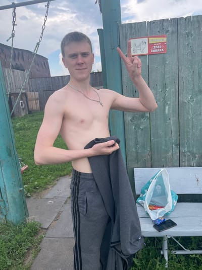 A young, shirtless Caucasian male with short, light brown hair and fair skin stands in front of a weathered, teal-painted wooden fence. He is wearing grey athletic pants with black stripes down the sides. He holds a dark grey garment in his left arm and extends his right arm, making a peace sign with two fingers. A sign on the fence reads "ОСТОРОЖНО ВО ДВОРЕ ЗЛАЯ СОБАКА" (Caution, beware of the fierce dog) with a drawing of a dog. To his left, a metal swing set with chains is visible, and behind him are wooden buildings and overgrown grass. To his right, a light blue wooden bench sits on the grass. On the bench are a plastic bag with items inside, a pack of cigarettes, and a black smartphone. The scene appears to be an outdoor, rural residential area, possibly a dacha or village in Nizhnii Bugrysh, Russia, given the context. The lighting suggests it is daytime, with a cloudy sky overhead.