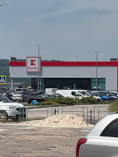 This media file captures an outdoor scene with a large retail building, likely a supermarket, in the background. The building features a white facade with a prominent red stripe running horizontally across its upper section. A large, square sign with the Kaufland logo, a stylized red "K" within a white square, is visible on the building. The word "Kaufland" is also written in white text on a red background next to the logo.
In front of the building, a parking lot is filled with various vehicles, including cars and vans. The vehicles are parked in rows, suggesting a typical shopping day. Some greenery, consisting of bushes and trees, lines the edge of the parking lot and the building. The ground of the parking lot appears to be paved, with some areas showing gravel or dirt.
A few streetlights or poles are visible in the scene, one with a small wind turbine-like attachment. The sky is overcast with a pale blue hue, indicating cloudy weather. The overall lighting suggests it is daytime, possibly late morning or afternoon.
A yellow and blue sign with a pictogram of a person walking is visible on a pole in the foreground, which could be a pedestrian crossing or directional sign. A white van is partially visible in the lower right corner of the image, suggesting the perspective is from someone in or near a vehicle.
The scene appears to be in Orhei, Moldova, as indicated by the provided location context. There are no people clearly visible in the image. The overall impression is a typical retail environment on a cloudy day.