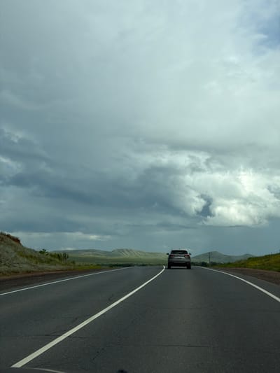 The image shows a dark grey asphalt road with white lane markings, traveling through a rural landscape under a cloudy sky. In the distance, a silver SUV is driving away from the viewer on the same road. The landscape on either side of the road is characterized by rolling hills covered in green grass and some scattered yellow wildflowers. On the left side of the road, there is a reddish-brown embankment with sparse vegetation. The sky is filled with dramatic, dark grey clouds, suggesting an impending storm or heavy overcast. The overall impression is one of an open, somewhat desolate highway journey through a vast natural setting. The location is identified as Verkhneozernoye, Russia.