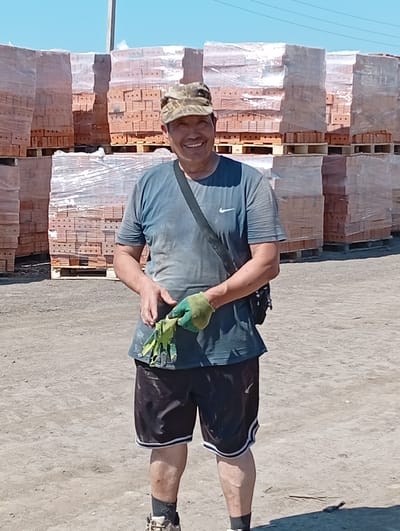 A man stands outdoors in an industrial area, possibly a brickyard, with stacks of bricks wrapped in plastic behind him. He is wearing a camouflage baseball cap, a blue t-shirt with a white Nike logo, black shorts with white trim, and work gloves. He is holding a bunch of green leafy material in his right hand, which is gloved. The ground is dirt and gravel, and the sky is clear and blue. There are no discernible identifying markers of the city of Қызылжар ауылдық округі, Kazakhstan within the image itself.