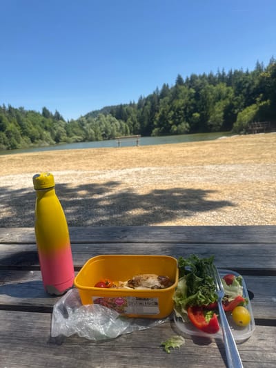 A bright yellow and pink water bottle stands upright on a weathered wooden picnic table. Next to it sits a yellow plastic food container holding a portion of cooked food, likely chicken, with a few cherry tomatoes. To the right of the container is a clear plastic container filled with a fresh salad, featuring lettuce, red bell pepper slices, and a small yellow tomato. A silver fork rests on top of the salad. A crumpled plastic bag is visible near the food container. The background of the image reveals a body of water, a sandy or gravelly shore, and a dense forest bordering the water. The sky is a clear, vibrant blue. The scene suggests an outdoor picnic at a lake or reservoir. The location in Coteaux du Lizon, France, implies a natural, possibly recreational setting. There are no people visible in the image.