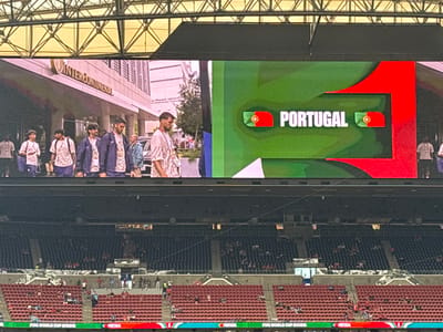 This is a photograph taken at a stadium, likely during a sporting event. The main focus of the image is a large LED screen displaying information related to the FIFA World Cup 2026. The screen shows the word "PORTUGAL" in large white lettering, accompanied by the Portuguese flag. A green and red graphic design frames the text and flag.
Below the screen, rows of empty stadium seating are visible, suggesting that the event has either not yet started or is in its early stages, or that the crowd is sparse. In the background, beyond the screen, a building with the "INTERCONTINENTAL" logo is visible, indicating an urban setting, possibly Houston, Texas, given the context provided.
In the foreground, partially obscured by the stadium structure, are several people. They appear to be walking or standing, and their attire is casual, suggesting they are spectators or staff. The lighting suggests it is daytime. The overall impression is one of anticipation for a major international sporting event. The text "FIFA WORLD CUP 2026" is also visible at the bottom of the screen.