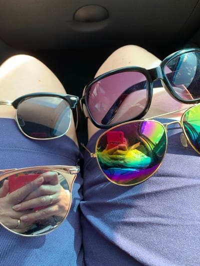 The image depicts a close-up view of a person's lap, covered by a textured blue fabric, likely clothing. Several pairs of sunglasses are arranged on the fabric. In the foreground, a pair of gold-rimmed sunglasses with mirrored, rainbow-colored lenses dominates the view. The reflection in these lenses shows a person holding a red smartphone and wearing rings on their fingers. Behind these, to the left, is a pair of black-rimmed sunglasses with dark lenses. To the right, another pair of black-rimmed sunglasses with purple-tinted lenses is visible. The background is dark and out of focus, suggesting an indoor or shadowed environment, possibly the interior of a vehicle. The image does not provide information about the specific location within Азерскі сельскі Савет, Belarus.