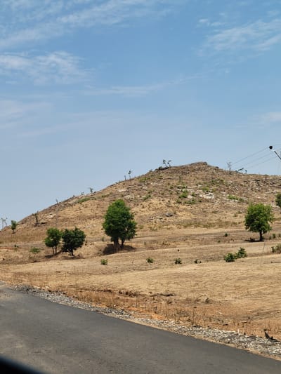 The image shows a roadside view of a dry, hilly landscape in Jama'a, Nigeria. The hillside is sparsely vegetated with brown grass and scattered trees. A paved road runs along the base of the hill. Power lines are visible along the top of the hill. The sky is light blue with some clouds.
