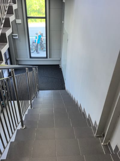 The image shows an interior staircase with a window looking out onto a street. Two children, a boy and a girl, are visible through the window, standing next to a bicycle. The boy is wearing a blue t-shirt and the girl is wearing a green cap and a blue t-shirt. The staircase is made of grey tiles and has a metal railing. The window is large and rectangular, with a black frame. The exterior scene outside the window is blurred, but trees and a street are visible. The overall impression is that of a modern building with a view of the outdoors. The location is the city of Zima Juzhnaja, Russia.
