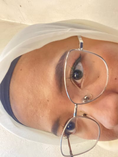 An overhead close-up shot shows a person's face wearing glasses. The person is wrapped in a white hijab and a dark headscarf is visible underneath. The person is looking down, and their eyes are visible through the glasses. The background is a textured off-white surface. There is no indication of the location being Kembang Harum, Indonesia.