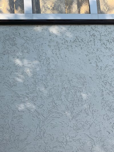 The image shows a light gray textured wall with a black trim running horizontally across the top. Above the trim is a glimpse of decorative fabric with a gold damask pattern, suggesting a window or awning. Shadows of foliage are cast across the textured surface of the wall, indicating an outdoor setting. The overall scene is static and lacks any people or explicit actions. The location is specified as Karmanovo, Russia.