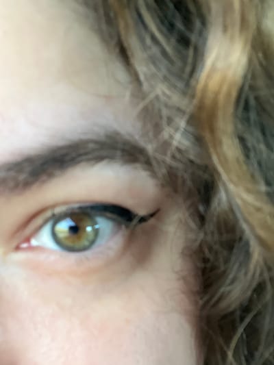 This is a close-up, slightly blurry, eye-level photograph of a person's eye, taken in Manolsko Konare, Bulgaria. The subject's eye is the central focus, with strands of light brown and blonde hair blurred in the upper portion of the frame. The eye is a hazel color, with a dark pupil and lighter brown and green tones in the iris. A reflection of what appears to be a window or bright light source is visible in the pupil. The skin around the eye is fair, with some reddish undertones visible on the eyelid and under the eye. The person's eyebrow is dark and appears to be well-defined. The lighting suggests it could be daytime, with the natural light illuminating the subject's face. No specific activity or event is clearly depicted, other than the subject's gaze, which is directed slightly to the left of the frame. There is no visible text in the image. The overall mood is intimate and personal, focusing on the detail of a single human eye.