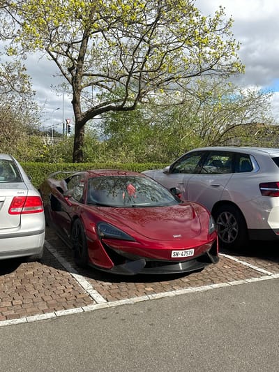 The image shows a red McLaren sports car parked between a white SUV and a silver sedan in a parking area adjacent to a brick-paved surface. The car has a license plate reading "SH-47579". In the background, there is a traffic light visible through the branches of a tree with budding yellow leaves. The environment suggests an urban setting, specifically in Schaffhausen, Switzerland, based on the license plate code "SH". The sky is partly cloudy.