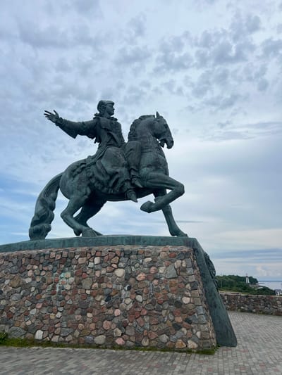 An equestrian statue of Peter the Great stands on a stone base in Baltiysk, Russia. The statue is made of bronze and depicts Peter the Great in a historical costume, pointing forward with his left hand. The horse is rearing up on its hind legs. The base of the statue is made of rough-hewn stones. In the background, there is a body of water, a lighthouse, and some trees. The sky is overcast with clouds.
