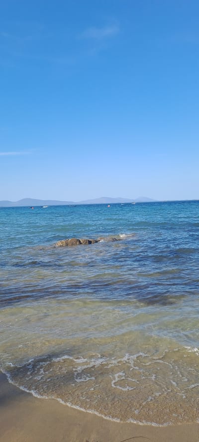 The image depicts a serene coastal scene under a clear blue sky. In the foreground, a sandy beach gently slopes into the calm, shallow water. The water is transparent, revealing the sandy bottom beneath, with small waves creating white foam as they reach the shore. Further out, the sea deepens to a rich blue, dotted with a few small boats and buoys. In the mid-ground, a cluster of rocks juts out of the water, partially submerged and weathered by the sea. In the distance, a line of low, hazy mountains forms the horizon. The overall impression is one of peace and natural beauty.